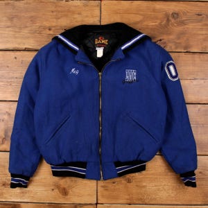 May include: A blue varsity jacket with a black collar and cuffs, detailed with white stripes. The jacket has a zip closure and a hood. The name "Meg" is embroidered on the chest, and the number "0" is on the sleeve.