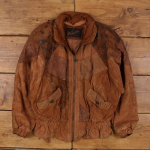 Puede incluir: Chaqueta de ante marrón con cremallera, dos bolsillos delanteros y cintura fruncida. La chaqueta presenta un diseño de patchwork con detalles en marrón oscuro y el nombre de la marca "Wimlit" en el cuello.