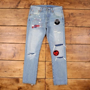 May include: Light blue denim jeans with distressed details, including rips and patches. Patches include a red heart, a circular design, and a design with red, white, and blue colours. The jeans are displayed on a wooden surface.