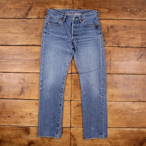 Könnte beinhalten: Hellblaue Jeans mit Knopfleiste, fünf Taschen und geradem Bein. Die Jeans sind an manchen Stellen verblichen und weisen Gebrauchsspuren auf. Das Kleidungsstück liegt flach auf einer Holzoberfläche.