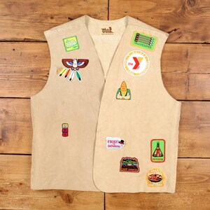 May include: Tan vest with various colorful patches, including a Native American design, a corn cob, and the YMCA logo. The vest has a simple, open front design and is likely vintage.