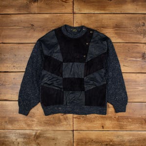 May include: A vintage sweater with a patchwork design. The front features a geometric pattern of black and dark gray panels. The sleeves are a speckled blue knit, and the collar is ribbed. Gold-colored buttons accent the shoulder.