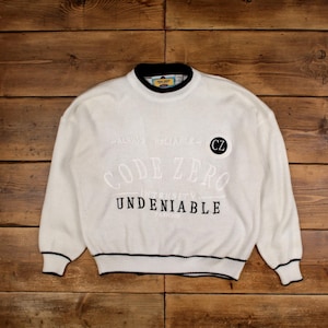 Peut inclure: Pull en tricot blanc avec bordures noires au col, aux poignets et à l'ourlet. Le pull porte l'inscription "CODE ZERO" en grandes lettres blanches, ainsi que les mots "ALWAYS RELIABLE" et "UNDENIABLE" en plus petit. Un écusson circulaire avec "CZ" est également visible.