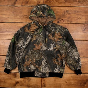 May include: A zip-up hooded jacket featuring a camouflage pattern of brown, green, and black forest foliage. The jacket includes a brown fleece-lined hood, long sleeves with black ribbed cuffs, and a black ribbed hem at the waist for a secure fit.