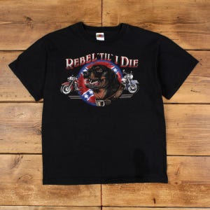 Peut inclure: T-shirt noir avec un motif graphique. Le motif représente un chien, des motos et le texte "Rebel 'Til I Die". Le graphisme comprend un emblème circulaire rouge, blanc et bleu avec un visage de chien et un drapeau confédéré, avec deux motos.