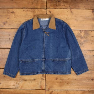 May include: A blue denim jacket with a brown corduroy collar. The jacket features a zip closure, two front pockets, and a leather pull tab. The jacket is displayed on a wooden surface.