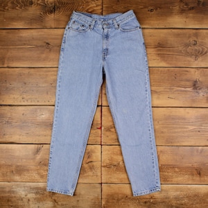 May include: Light blue denim jeans with a classic five-pocket design. The jeans have a straight leg cut and a high waist. The denim has a faded appearance, with subtle wear marks. The jeans are laid flat on a wooden surface.
