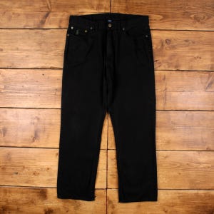 May include: Black denim jeans with a classic five-pocket design. The jeans feature a button and zip closure, belt loops, and a small embroidered logo on the front pocket. The jeans are displayed on a wooden surface.