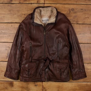 Peut inclure: Veste en cuir marron avec doublure en fausse fourrure beige. La veste a une fermeture éclair, deux poches de poitrine, deux poches inférieures et une ceinture. La marque "MARTANI" est visible.