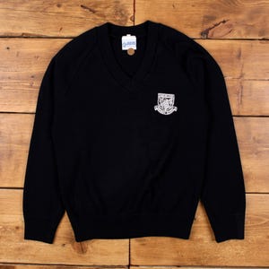 Peut inclure: Pull noir à col en V avec un écusson blanc sur la poitrine. Le pull a des manches longues et des bordures côtelées. L'écusson présente un motif de bouclier avec du texte. Le pull est fait d'une matière douce.