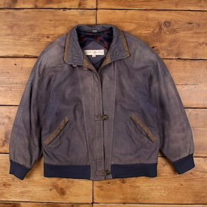 Könnte beinhalten: Vintage blaue Lederjacke im Bomber-Stil. Die Jacke hat einen Kragen, zwei Vordertaschen und einen Knopfverschluss. Die Manschetten und der Saum sind dunkelblau. Die Jacke wird auf einer Holzoberfläche präsentiert.