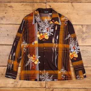 May include: A brown long-sleeved shirt with a plaid pattern and floral print. The shirt has a button-up front and a collar. The print features orange, yellow, and white flowers and leaves.
