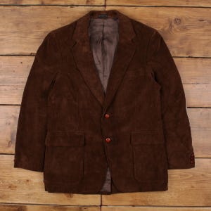 Puede incluir: Blazer de pana marrón con cierre de dos botones y dos bolsillos delanteros. El blazer tiene solapas con muescas y detalles de botones en las mangas. La chaqueta se muestra sobre una superficie de madera.