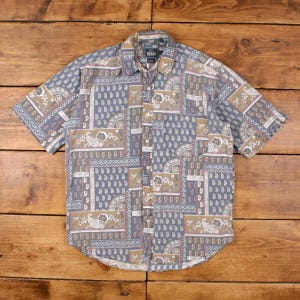May include: Short-sleeved button-down shirt with a patchwork design. The shirt features a paisley pattern in shades of blue, tan, and white. The shirt has a collared neckline and a button-up front. The shirt is made of a lightweight fabric.