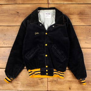 May include: A black corduroy varsity jacket featuring a quilted light grey interior lining, a fold-down collar, and yellow snap buttons. The name John is embroidered in yellow on the chest. The cuffs and hem have yellow and black striped trim.