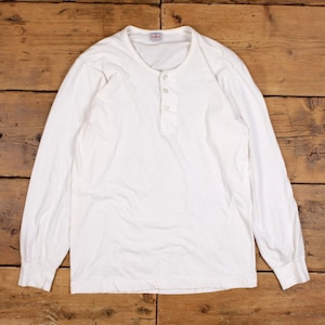 Puede incluir: Una camiseta henley blanca de manga larga con cuello redondo y una tapeta de tres botones. La prenda está confeccionada en tejido blanco liso e incluye una pequeña etiqueta rectangular de marca con el texto R.A.W. en el interior del cuello.