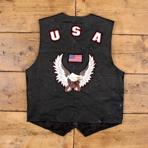 May include: Black leather vest with a patriotic theme. Features a large eagle patch with outstretched wings, a small American flag, and the letters "USA" in red and white. The vest has a button closure and is displayed on a wooden surface.