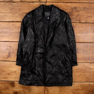 May include: A black leather jacket with a long, knee-length design. The jacket has a notched collar, a zipper closure, and the text "GENUINE LEATHER" on a tag. The jacket is displayed on a wooden surface.