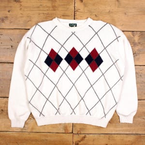 Peut inclure: Pull en maille blanc à col rond avec motif losanges. Le pull présente un motif de losanges noirs sur fond blanc, avec trois losanges centraux rouges et bleu marine. Fabriqué dans une matière douce et confortable.