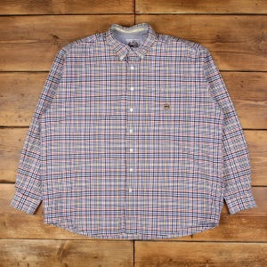 Puede incluir: Una camisa abotonada de manga larga con un estampado a cuadros en azul, rojo, amarillo y blanco. La camisa tiene cuello abotonado y un pequeño emblema bordado en el pecho. La camisa está tendida sobre una superficie de madera.