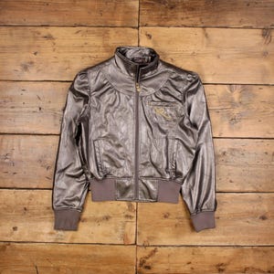 May include: A metallic grey bomber jacket with a high collar, zip-up front, and two front pockets. The jacket has a brown ribbed hem and cuffs. A gold logo is embroidered on the chest. The jacket is displayed on a wooden surface.