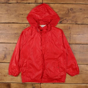 May include: A vibrant red hooded jacket with a zip and two front pockets. The jacket is made of a lightweight material and is laid flat on a wooden surface. The hood is attached and the jacket appears to be vintage.