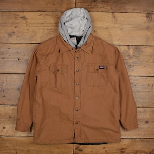 May include: A tan long-sleeve button-up jacket featuring a light gray jersey hood, a front button closure, and two chest pockets with buttoned flaps. A small black rectangular brand label is stitched onto the upper right chest pocket.
