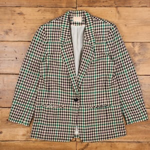 May include: A black, green, and cream checked blazer. The jacket features a single-button closure, two front pockets, and a light blue lining. The blazer is displayed on a wooden surface.