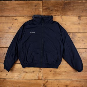 Op de afbeelding: Een marineblauwe Columbia Sportswear jas met een volledige ritssluiting aan de voorzijde, een hoge opstaande kraag en lange mouwen met elastische boorden. Het merklogo is in het wit geborduurd op de rechterbovenkant van de borst.
