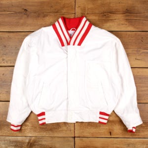 May include: White bomber jacket featuring a red and white striped collar and cuffs. The jacket has a front pocket and a button closure. The jacket is made of a smooth material. The jacket is likely vintage or retro.