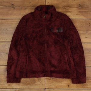 May include: A burgundy fleece jacket with a full-length zipper and a high collar. The jacket features a small plaid pocket on the chest and a plaid lining inside the collar. The soft, textured material suggests warmth and comfort.