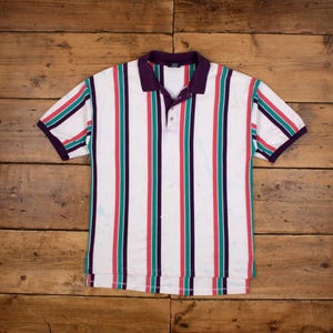 May include: White polo shirt with vertical stripes in red, teal, and purple. The shirt has a purple collar and sleeve trim. The shirt is made of a soft material and has a button-up collar.