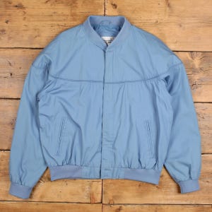 May include: A pale blue bomber jacket with a full front zip. The jacket has a ribbed collar, cuffs, and hem. It features two front pockets and a light blue lining. The jacket is made of a lightweight material.