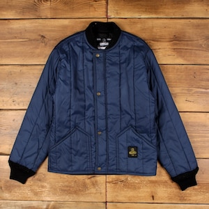 May include: A navy blue quilted jacket featuring a black ribbed collar and cuffs, a front button closure with metallic buttons, and two front pockets. A small black rectangular patch with yellow text and a graphic is sewn onto the lower right pocket.