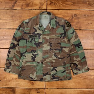 Op de afbeelding: Camouflage jas met meerdere zakken en de naam "MENENDEZ" geborduurd boven de linker borstzak. De jas heeft een knoopsluiting en een militair design, met groene, bruine en zwarte kleuren.