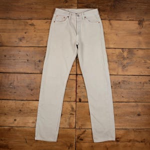 May include: Light beige denim jeans with a classic five-pocket design. The pants are straight-legged and have a button and zipper closure. The jeans are laid flat on a wooden surface.