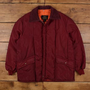 May include: A burgundy quilted jacket with a corduroy collar and orange lining. The jacket has two front pockets and a zipper closure. The cuffs are ribbed. The jacket is displayed on a wooden surface.