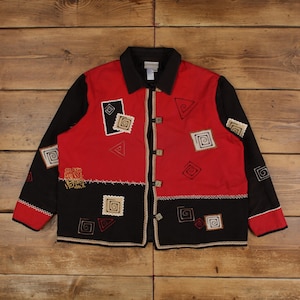 Puede incluir: Una chaqueta de manga larga con botones y diseño de bloques de color en rojo y negro. La prenda incluye patrones bordados de espirales cuadradas y triangulares, un cuello negro y cierres decorativos de palanca en la parte delantera.