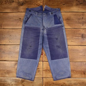 May include: Light blue denim work pants with darker blue patches on the knees and back pockets. The pants have a button fly and a relaxed fit. The denim has a worn, distressed appearance.