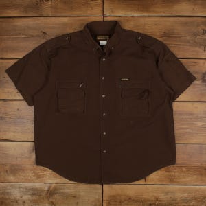 Peut inclure: Une chemise marron à manches courtes avec deux poches avant et des boutons. La chemise a un col et des épaulettes sur les épaules. Le tissu semble être un coton ou un mélange durable, adapté aux activités en plein air.