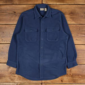 Puede incluir: Una camisa de forro polar azul marino con dos bolsillos en el pecho y una botonadura. La camisa tiene cuello y mangas largas. La etiqueta de la marca es visible en el cuello. La camisa se muestra sobre una superficie de madera.