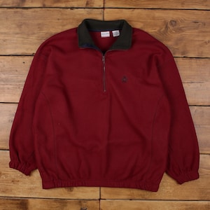 May include: A burgundy fleece pullover with a dark green collar and a quarter-zip closure. The long-sleeved top features a small embroidered emblem on the chest and elastic cuffs. The garment is displayed on a wooden surface.