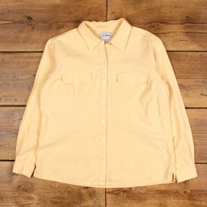 May include: A yellow long-sleeved button-down shirt with a small checkered pattern. The shirt has two chest pockets and a collar. The brand label "L.L.Bean" is visible on the collar.