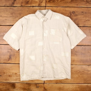 May include: A light beige short-sleeved shirt with a collared neckline and a single chest pocket. The shirt features rectangular patches of a lighter shade of beige. The shirt is made of a lightweight fabric.
