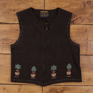 May include: A dark brown wool vest with a full zipper closure. The vest features embroidered flower pot designs along the bottom. The Woolrich label is visible at the neckline. The vest has a round neckline and a sleeveless design.
