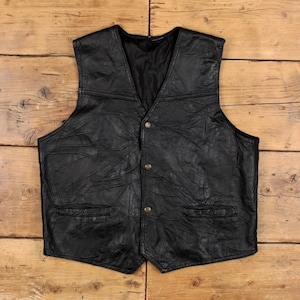 May include: A black leather vest with a V-neck, two front pockets, and three gold-coloured buttons. The vest has a textured, patchwork design and a black lining. The garment is displayed on a wooden surface.