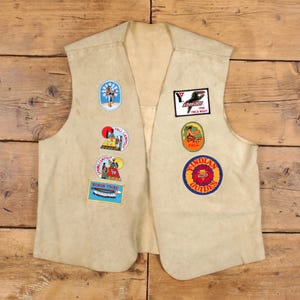 May include: A beige vest featuring several embroidered patches. Patches include a YMCA logo, "Indian Guides", and camping-themed designs. The vest is made of a soft material and has a V-neck.