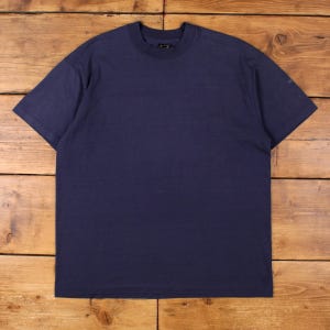 Könnte beinhalten: Ein marineblaues T-Shirt mit kurzen Ärmeln. Das Shirt hat einen Rundhalsausschnitt und eine lockere Passform. Der Stoff scheint ein weiches, einfarbiges Material zu sein. Das Shirt wird auf einer Holzoberfläche präsentiert.