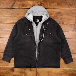 May include: A black button-up jacket featuring a built-in heather gray hooded sweatshirt insert. The jacket has a quilted interior lining, two chest pockets with snap closures, and a Dickies brand label on the interior and chest pocket.