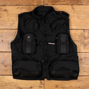 May include: A black utility vest featuring a central zipper closure, a high collar, and multiple storage pockets. The front includes two upper pockets with quilted panels and two large lower pockets with quilted bases. A Tamrac logo tag is visible.